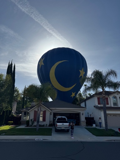 a photo of a house on a suburban street, and behind it, a huge hot air balloon large enough to carry 13 people and block the viewer's view of the morning sun