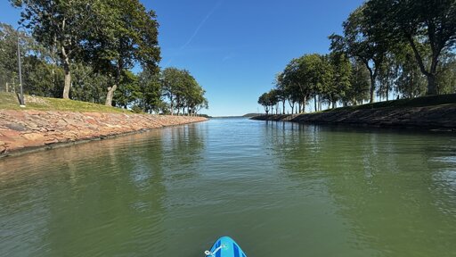 Photo from a channel with trees growing on both sides and blue skies ahead.