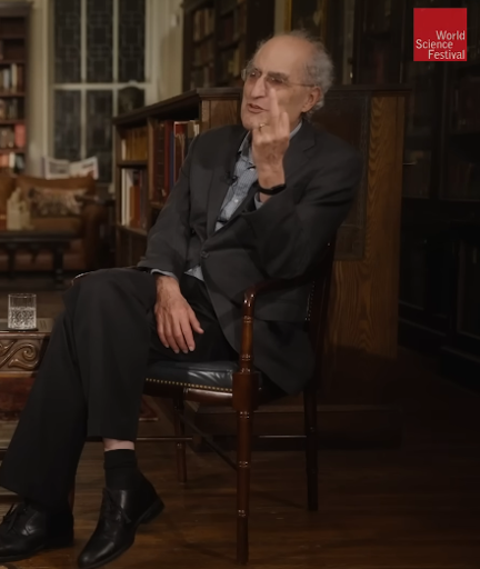 Ed Witten sitting in a chair holding up the middle finger of his left hand