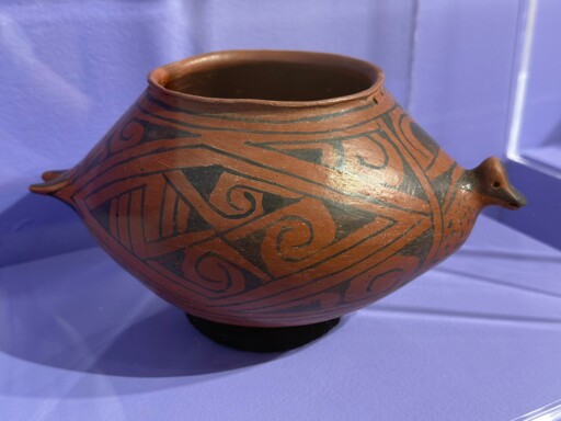 A reddish-brown, short and squat earthenware vessel with a decorative duck head on the front and a duck tail on the back. Swirling patterns are formed within the geometric diamond and triangle shapes that cover the vessel. The area right under the duck’s head is spotted like the chests of some female ducks 