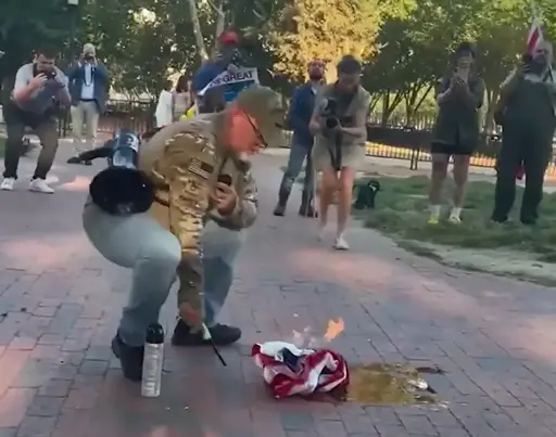 Guy in military clothing with an american flag on his shoulder burns an american flag in front of the white house while being filmed by bystanders